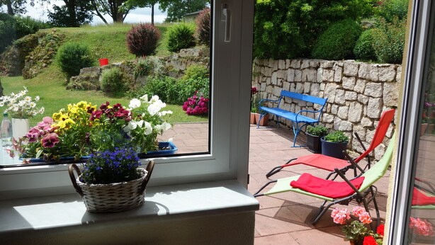 A window with a view of a stone wall, a bench, and a basket of flowers.