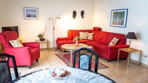 A living room with a red sofa, a round coffee table, and a dining table with chairs. There are two side tables, a floor lamp, and wall art.