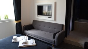 A modern hotel room with a grey sofa, a flat-screen TV mounted on the wall, and a bed with neatly folded white towels.