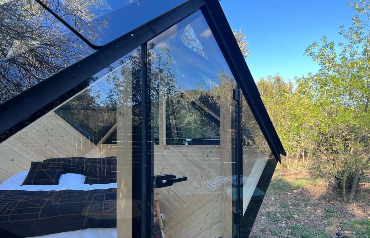A modern, glass-enclosed room with a bed and a view of the surrounding trees.