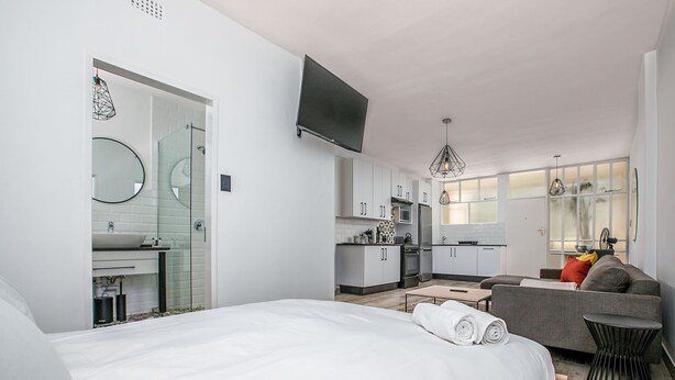 A modern hotel room with a large bed, a sofa, a dining table, and a kitchen area.