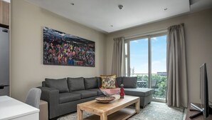 A modern living room with a grey sofa, a wooden coffee table, a large painting, and a view of the outdoors through a large window.