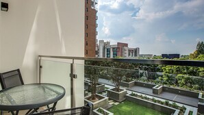 A balcony with a glass table and chairs, overlooking a landscaped garden and buildings in the background.