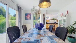 Een eethoek met een tafel, overdekt met een tafelkleed met een nautisch thema, omringd door rieten stoelen, een hanglamp en uitzicht op de omgeving.