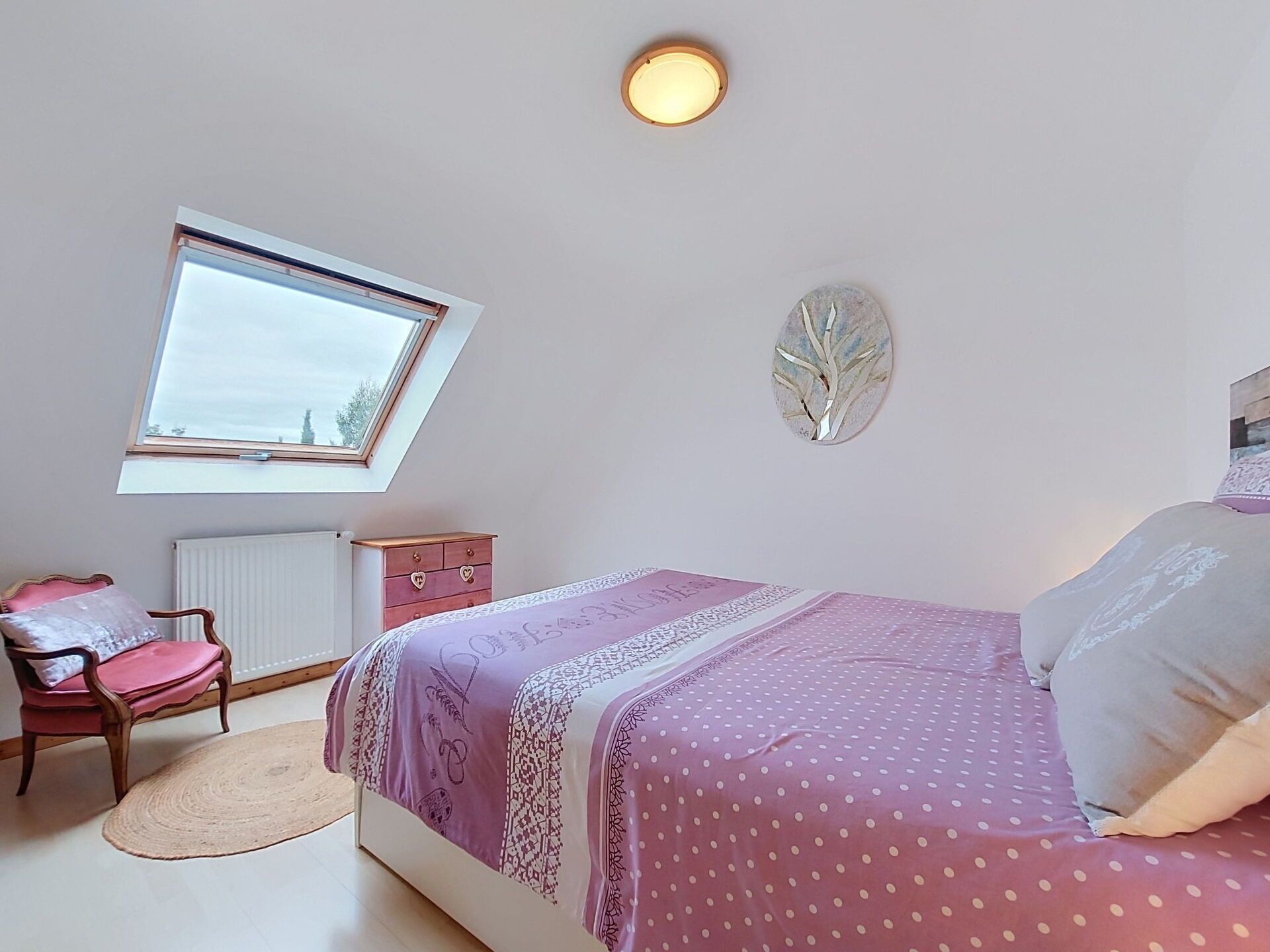 Een slaapkamer met een bed, een dressoir, een stoel, een radiator, een raam en een wanddecoratie.