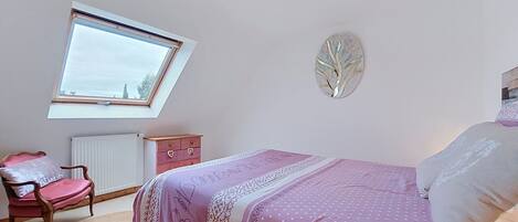 Een slaapkamer met een bed, een dressoir, een stoel, een radiator, een raam en een wanddecoratie.