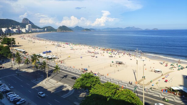 A beach with a road in the foreground and buildings in the background. - TC1001 Vista Perfeita de Copacabana (Rio de Janeiro)