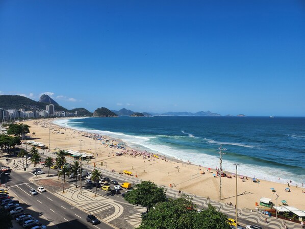 A coastal cityscape with a wide beach, clear blue sky, and distant mountains.