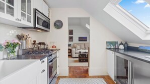 A modern kitchen with white cabinets, a microwave, and a stove. There is a skylight above the kitchen, and a living area with a couch and a painting visible through an open doorway.