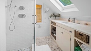 A modern bathroom with a glass-enclosed shower, a skylight, a white sink, and a beige vanity.