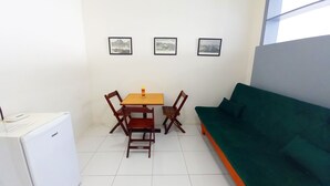 A small, clean room with a white refrigerator, a wooden table with two chairs, a green sofa, and three framed pictures on the wall. - TC1203 Janela para o Mar (Rio de Janeiro)
