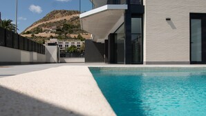 Un área de piscina moderna con una piscina azul cristalina, un deck blanco con textura y un edificio con grandes ventanales y techo plano.