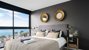 Un dormitorio moderno con una cama grande, dos espejos adosados a la pared, una mesita de noche con lámpara y vistas al mar.