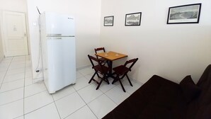 Un espacio habitable pequeño con un refrigerador blanco, una mesa de comedor de madera con cuatro sillas y un sofá negro. Hay tres cuadros enmarcados en la pared.
