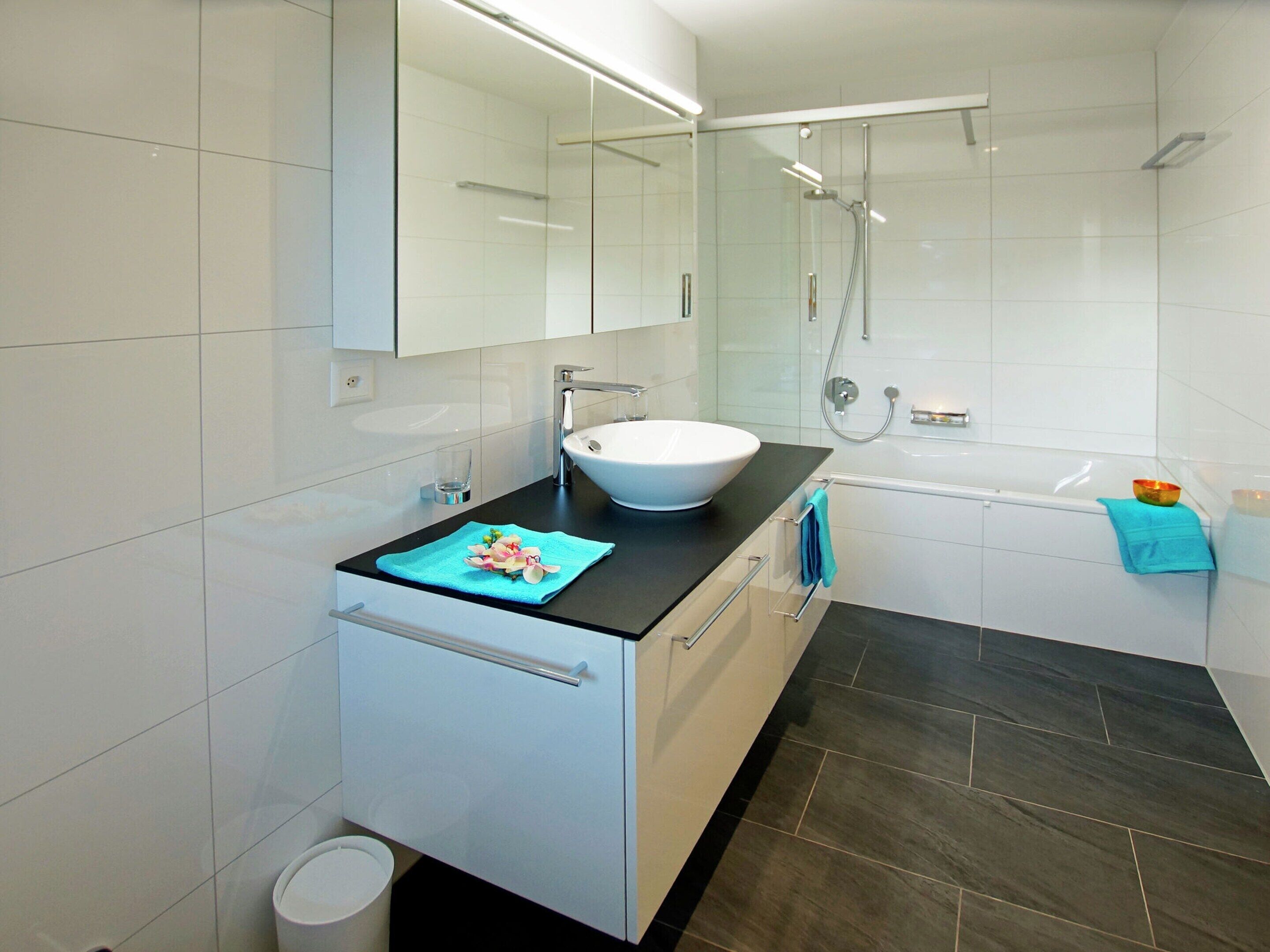 Ein modernes Badezimmer mit einem weißen Waschbecken, einer dunklen Arbeitsplatte und einer Duschkabine.