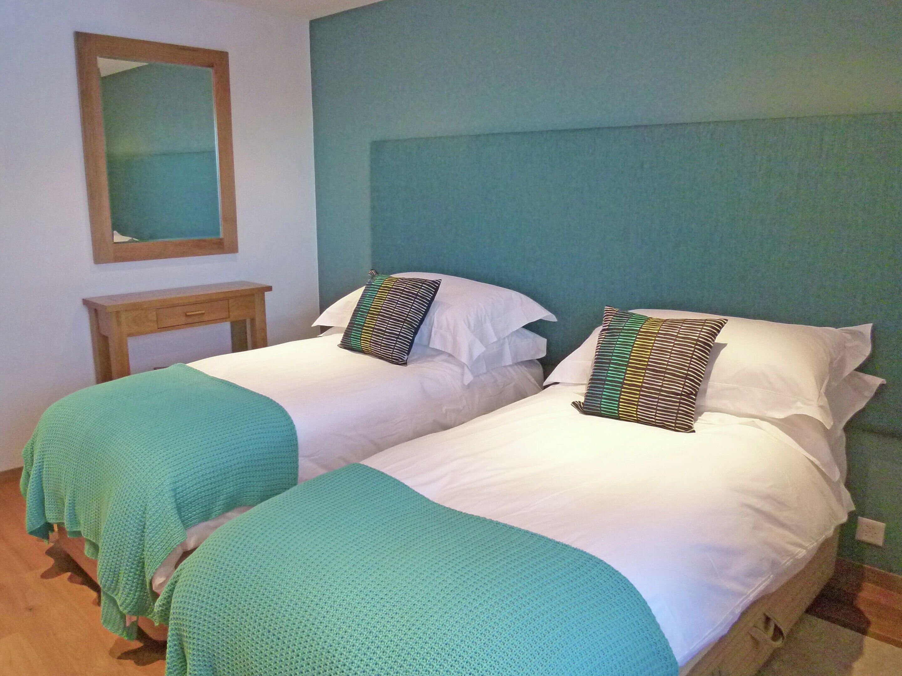 A twin room with two single beds, each with a green blanket and patterned pillows. A wooden nightstand and a mirror are visible.