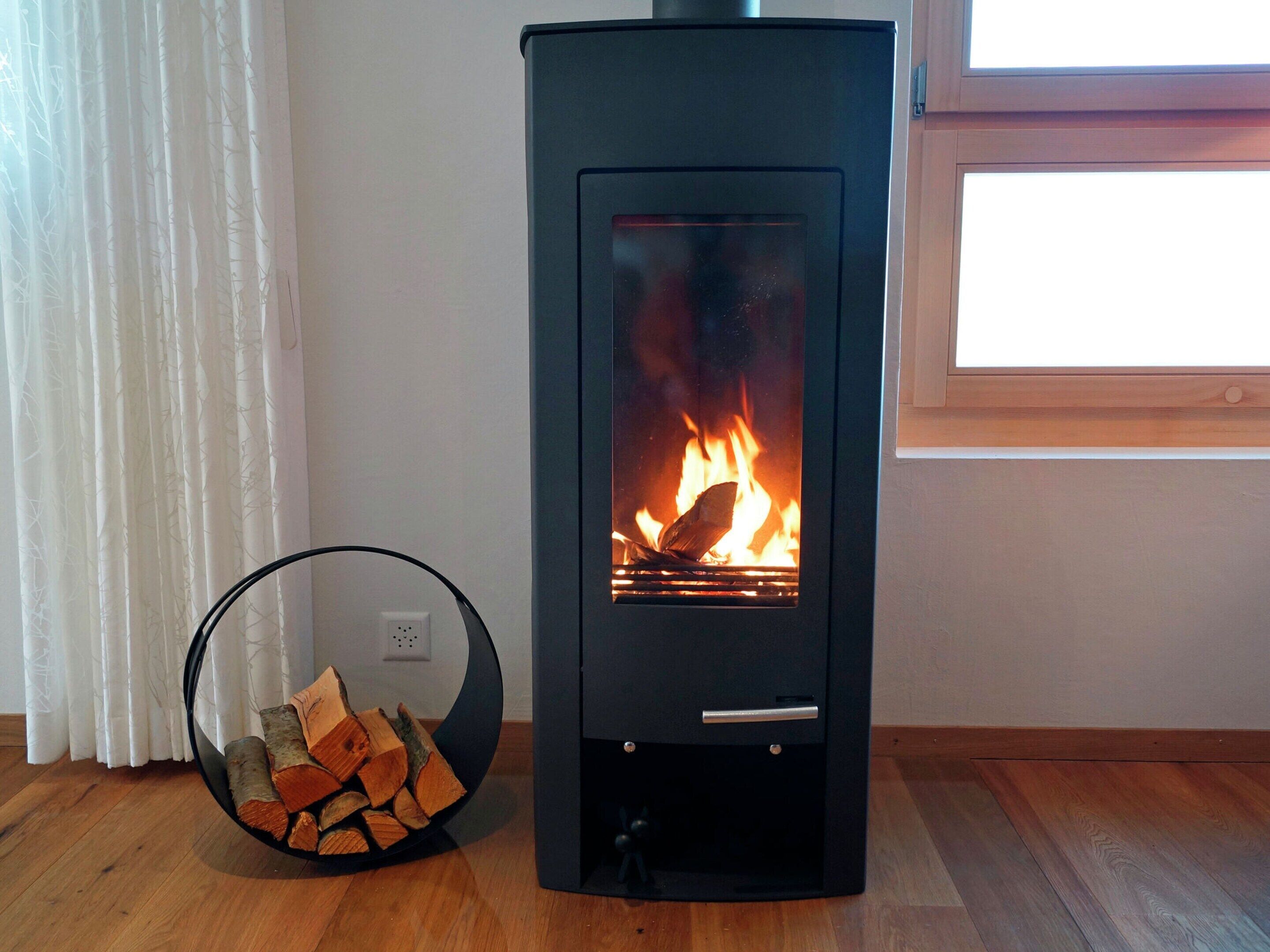 Un poêle à bois moderne doté d’une porte en verre, d’une poignée en métal noir et d’une petite étagère en dessous. Une pile de bois de chauffage est posée sur le sol à côté.