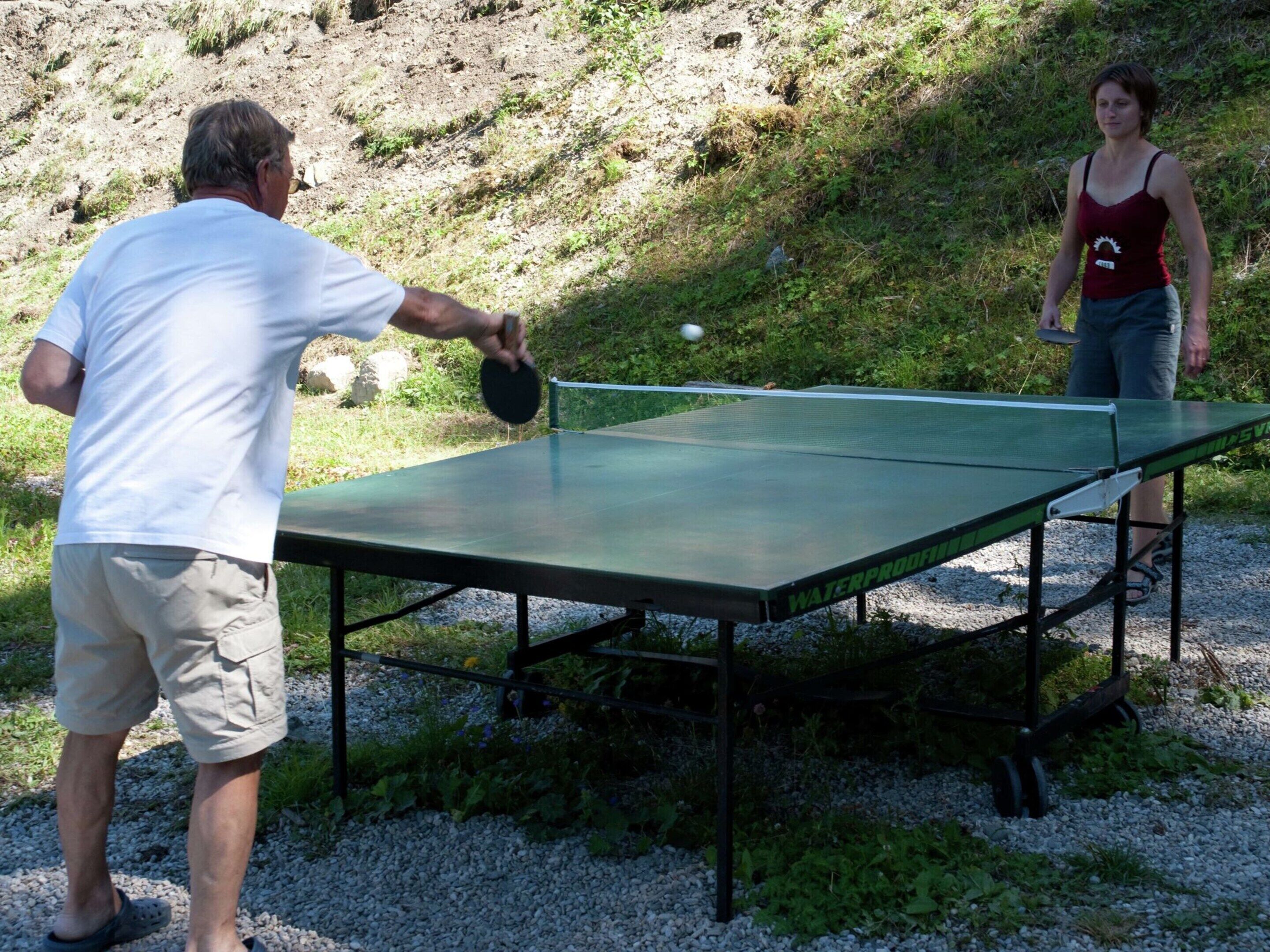A man and a woman playing table tennis outdoors on a green table with a net.