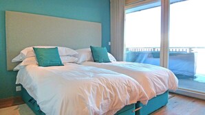 Cama doble con ropa de cama blanca y verde azulada, cabecera y vista a un balcón a través de un cristalero corredizo.