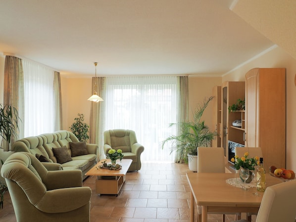 A living room with a sofa, armchair, coffee table, and a dining table with a vase of flowers. - Modernes Apartment in Kuehlungsborn mit Garten (Kuehlungsborn)