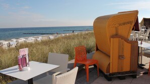 Ein Sitzbereich im Freien mit Korbstühlen und Tischen, mit Blick auf einen Strand und einen klaren Himmel.
