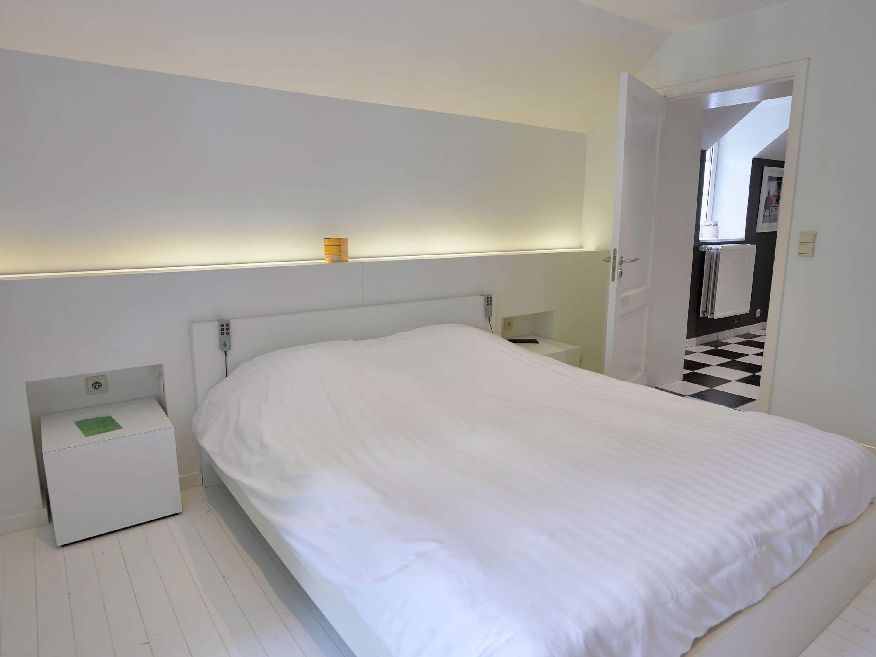 Une chambre à coucher avec un lit blanc, une table de chevet, une commode et une lampe fixée au mur.