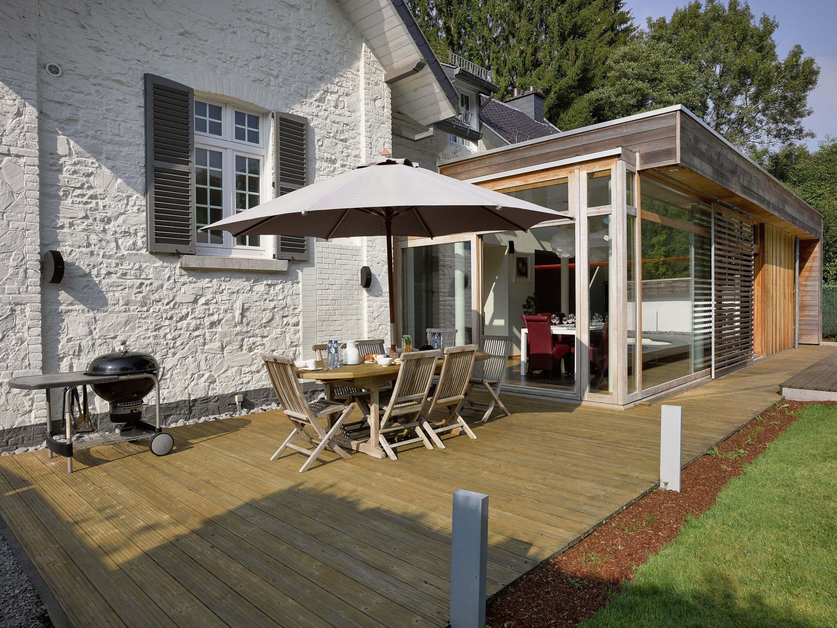 Un patio moderno al aire libre con una mesa y sillas de madera, una parrilla y un área de comedor con paredes de vidrio.