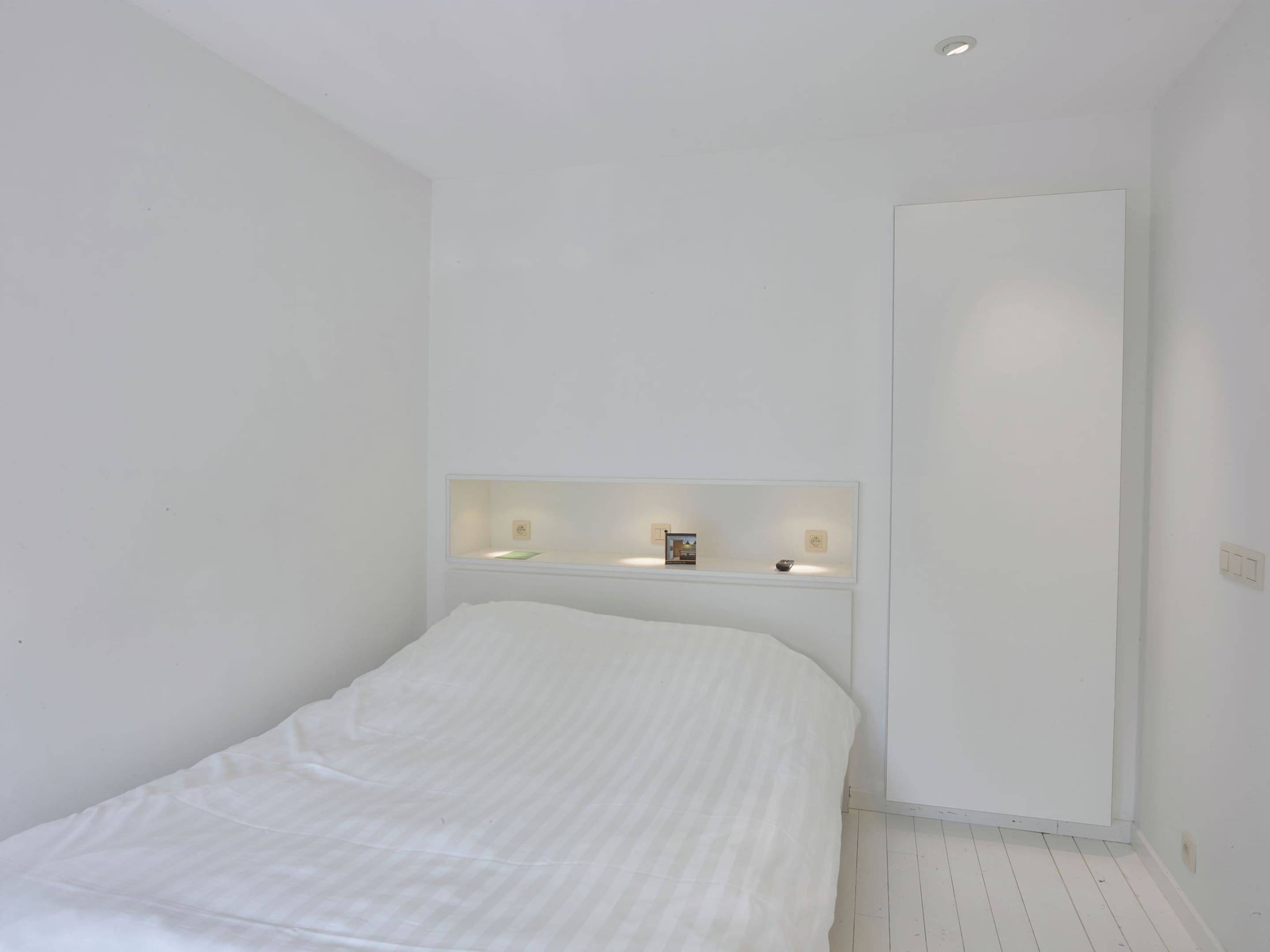 Une chambre à coucher au style minimaliste, avec un lit, une étagère intégrée agrémentée d’objets décoratifs et une grande armoire.