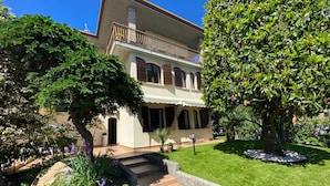 A two-story building with a balcony, a stone staircase leading to the entrance, and a well-maintained garden with various plants and a large tree. - Seclusive Apartment in Sirmione near Lake (Sirmione)
