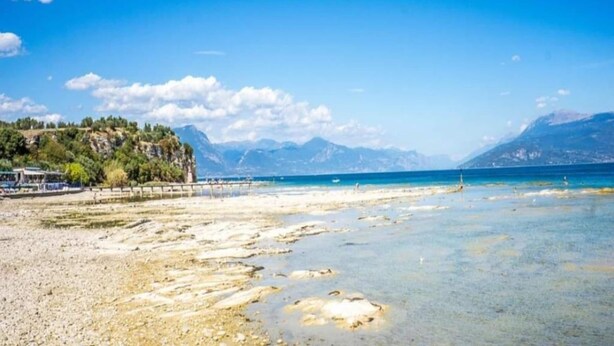 A beach with clear water, a rocky shoreline, and mountains in the background. - Seclusive Apartment in Sirmione near Lake (Sirmione)