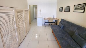 A small, clean room with a grey sofa, a desk with a chair, a small fridge, and a ceiling fan. - TC801 Charmoso Studio na Areia de Copacabana (Rio de Janeiro)