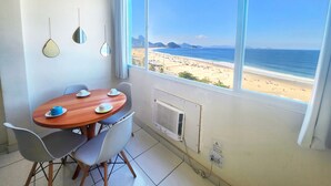 A room with a large window overlooking a beach, a dining table with chairs, and a radiator. - TC801 Charmoso Studio na Areia de Copacabana (Rio de Janeiro)