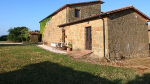Um edifício de pedra com telhado em telhas, um pequeno alpendre e uma mesa com cadeiras na parte externa.