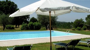 Piscina com guarda-sol branco, duas espreguiçadeiras e área gramada com árvores ao fundo.