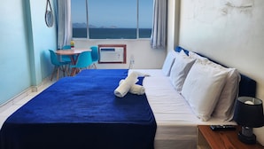 A hotel room with a bed, a table with chairs, a window with a view of the sea, and an air conditioning unit. - TC802 Vistão Praia de Copa (Rio de Janeiro)