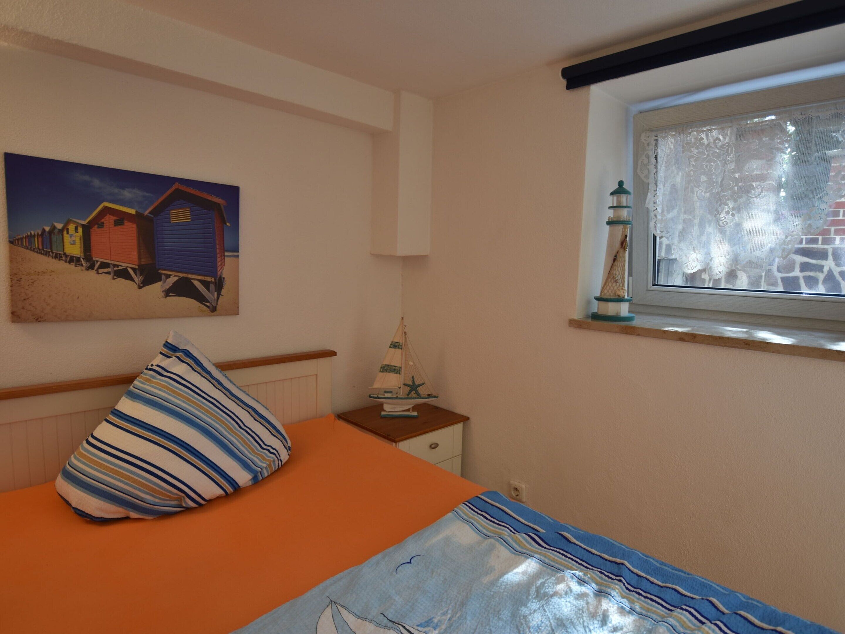 A bedroom with a bed, a striped pillow, a small table with a ship model, a painting of beach houses, and a window with a view of a brick wall.