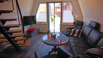 Ein modernes Wohnzimmer mit einem schwarzen Ledersofa, einem Holztisch und einem Fernseher auf einem Standfuß. Eine Holztreppe führt zu einer oberen Etage.