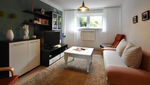 Ein Wohnzimmer mit Fernseher, Sofa, Couchtisch und einem Fenster mit Blick auf Grünflächen.