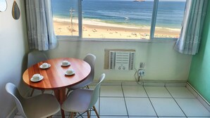 A room with a large window overlooking a beach, a round wooden table with chairs, and a wall-mounted air conditioning unit. - TC803 Vistão Av. Atlântica (Rio de Janeiro)