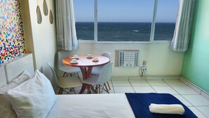 A room with a bed, a table with four cups, a window with a view of the sea, and a colorful wall decoration. - TC803 Vistão Av. Atlântica (Rio de Janeiro)