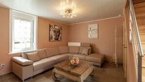 Un salón con un sofá seccional color beige, una mesa de centro de vidrio con flores, un cuadro de Buda y un radiador.
