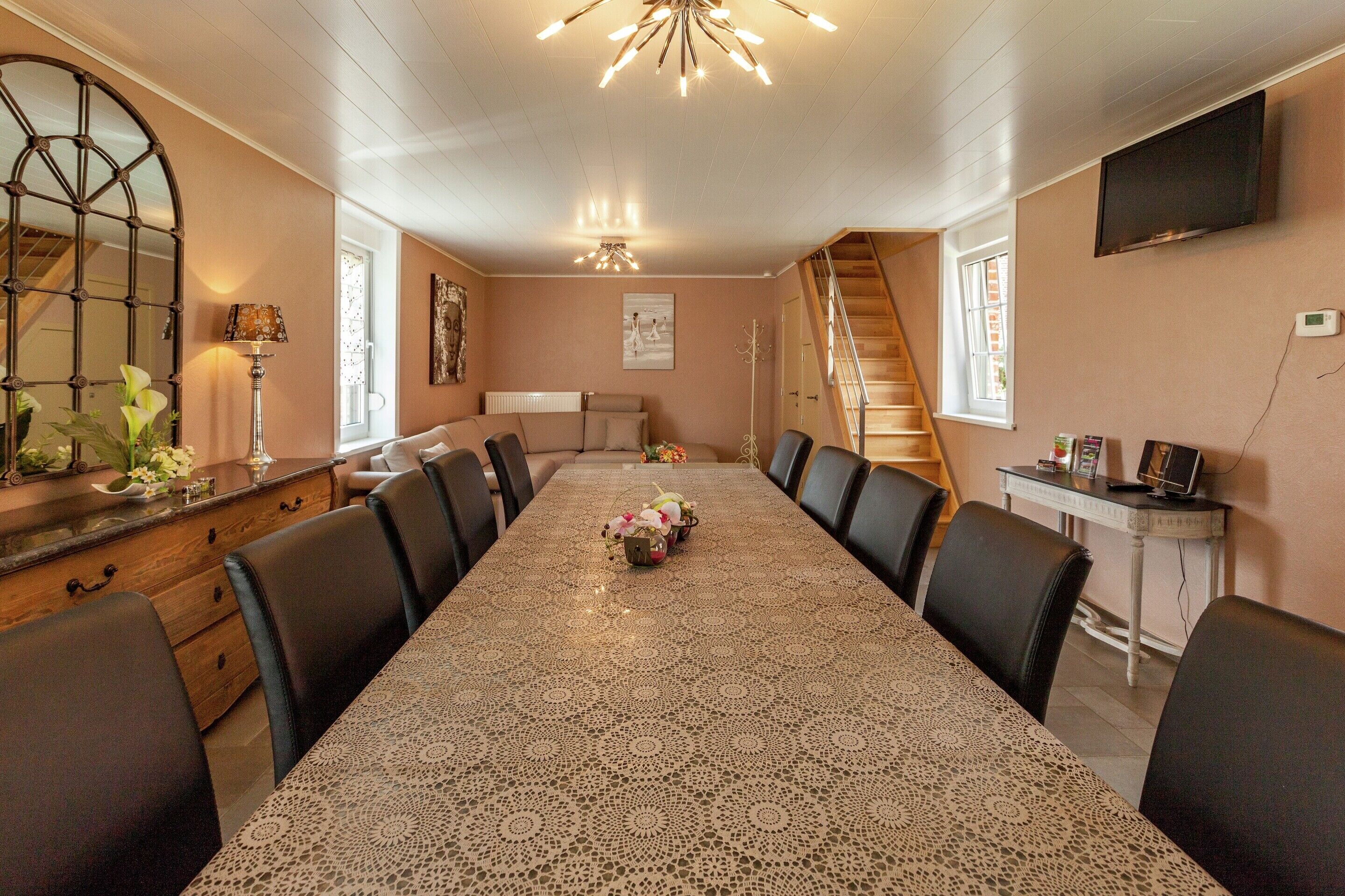 A dining room with a long table, chairs, a chandelier, a mirror, and a staircase.