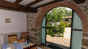 A dining area with a table set for two, a wicker chair, a stone wall, and a view of a courtyard with a barbecue grill. - Holiday House with Pool & Large Garden Overlooking Lake near Tuscany (Lisciano Niccone)