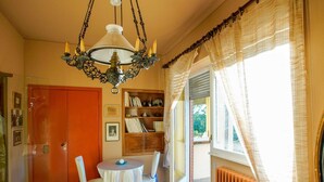Una habitación con una araña, una mesa redonda con sillas, una estantería y vistas a través de una ventana con cortinas.