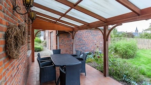 Een overdekt terras met een bakstenen muur, een tafel en stoelen, en een tuin en gebouwen op de achtergrond.