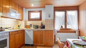 Ett kompakt kök med träskåp, en mikrovågsugn, en spis och en diskmaskin. Det finns ett fönster med gardiner och utsikt över grönområden utanför.