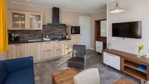 Eine moderne Küche mit Holzausstattung, einem schwarzen Herd und einer gefliesten Spritzschutzwand. Es gibt einen Essbereich mit einem Holztisch und Stühlen, sowie ein an der Wand montierter Fernseher.