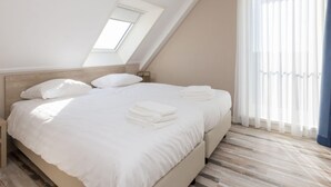 Ein modernes Schlafzimmer mit schrägen Wänden, einem Oberlicht, einem großen Bett mit weißer Bettwäsche und Blick ins Freie durch transparente Vorhänge.