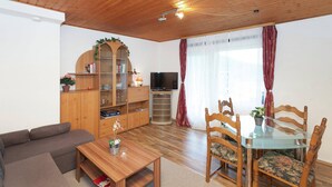 A living room with a wooden ceiling, a sofa, a glass dining table with chairs, a TV, and a wooden cabinet with shelves. - Apartment With Meadow View in Wildemann (Clausthal-Zellerfeld)