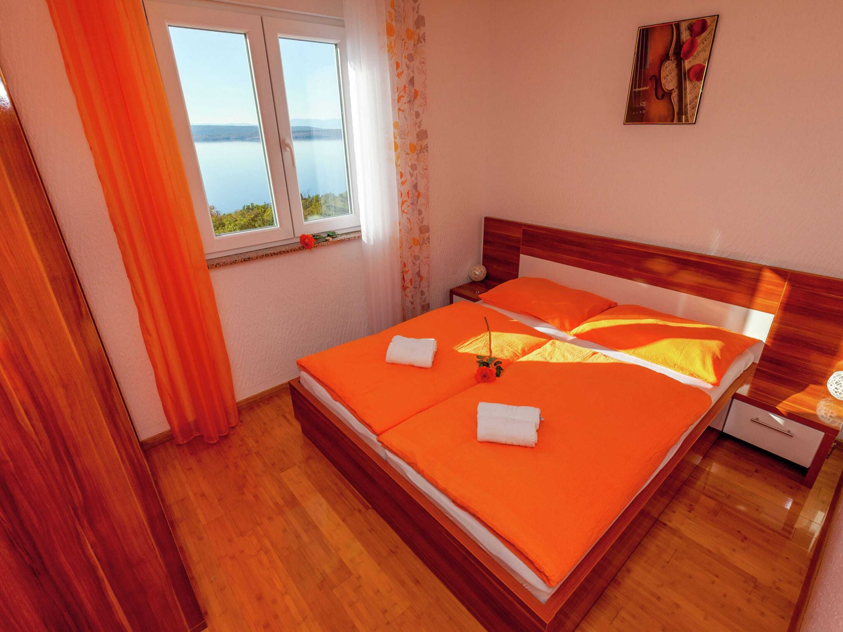 A hotel room with a wooden bed, orange bedding, a window with a view of the water, and a small table with a vase.